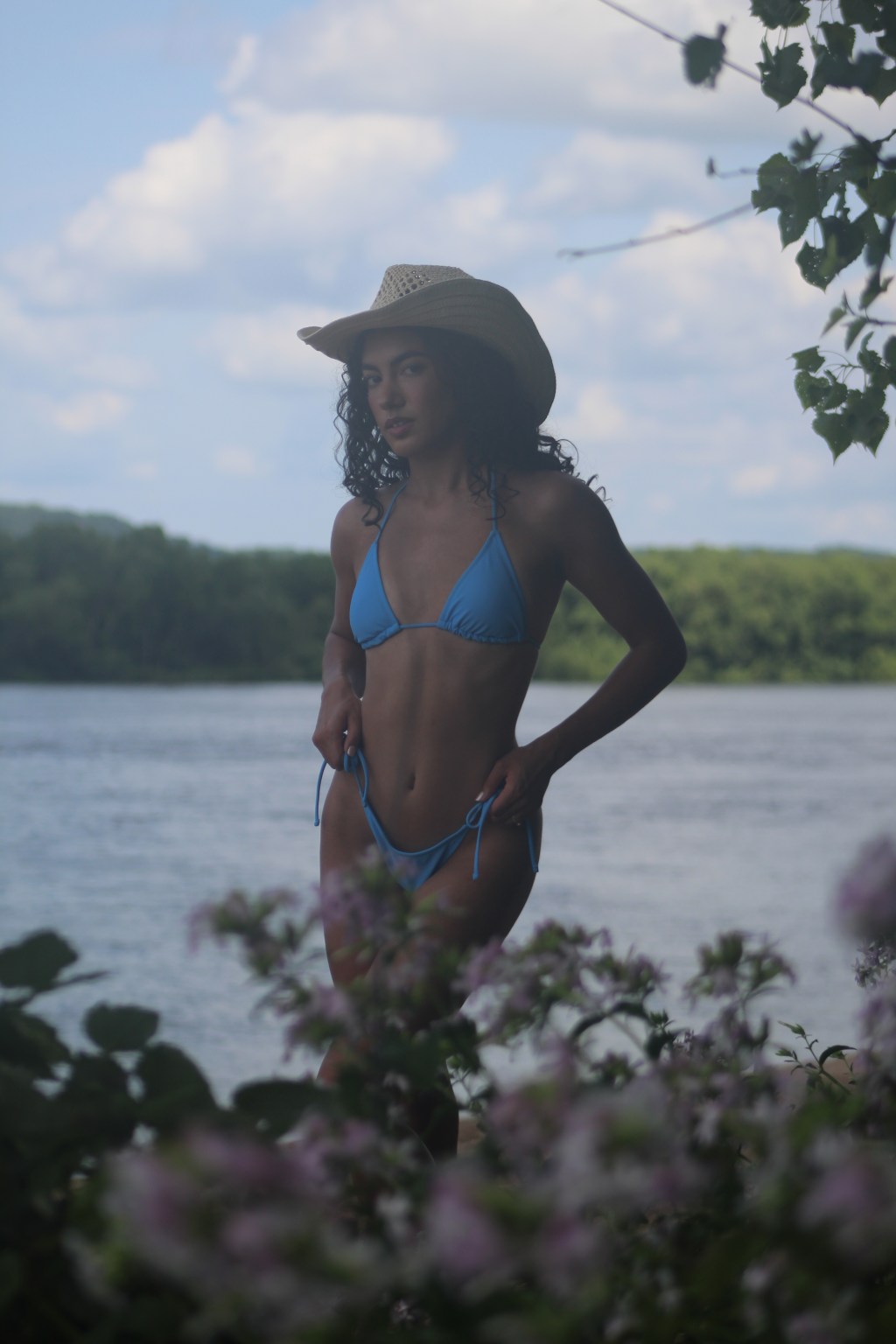 Model Erica posing outdoors by the river, wearing a vibrant blue bikini and a cowboy hat. She smiles confidently in natural sunlight, with water and greenery in the background, capturing playful summer and cowgirl vibes.