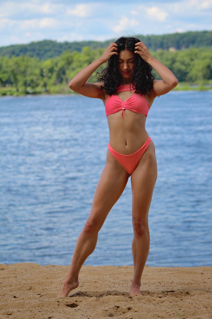 Model Erica posing outdoors by the river in a bright pink bikini and cowboy hat, enjoying the sunshine with playful, confident energy against a scenic natural backdrop.