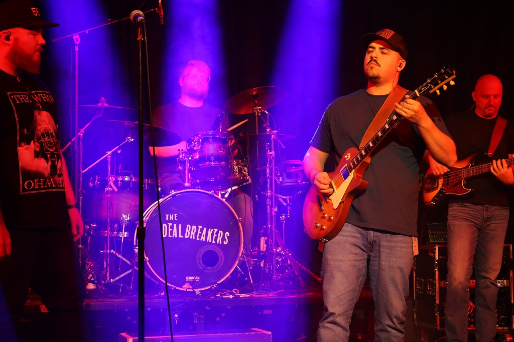 a band playing live on a stage. 3 prominent stage lights shine on a drum kit with the label on the bass that reads "the dealbreakers"